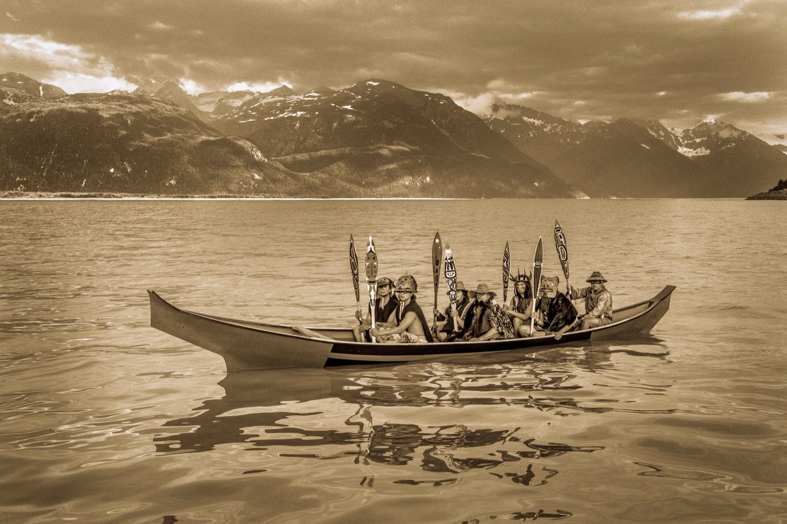 A carved war canoe of paddlers crossing still water below mountains